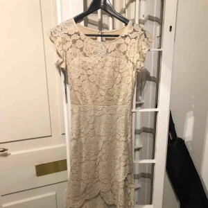  - Vacker beige spetsklänning som är kortare fram & längre bak.  Klänningen har vaddering i byst. Finns även dragkedja i rygg. Klänningen har ett fastsytt underlinne. Storlek S/M - passar en small eller en liten medium. Använd en gång.