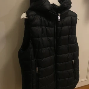 Svart väst MANGO  - En svart väst från MANGO i storlek L. Avtagbar luva. Silvriga detaljer. Västen är i bra skick. Köpt för 349kr. 