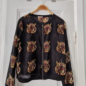 Tunn blus/jacka i polyester. Svart/mönst - Jättefin svart tunn blusaktig jacka i polyester. Mexikanskt märke. Dragkedja i guld. Mönster med tigrar mot svart bakgrund. Mjuk och skön, tycker om den men använder inte tillräckligt ofta 🙃. S/M.