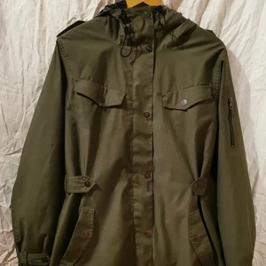 Everest : Grön jacka med huva - Gummisnodden som ska dra åt huvan är lite nött. Fyra fickor fram, en innerficka med nät och en "gömd" ficka vid dragkedjan där man kan ha grejer som man lätt vill komma åt utan att behöva öppna jackan. Snodd längst ner som man kan dra åt.