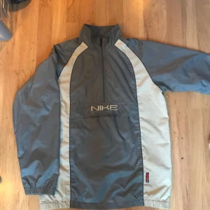  - Grå vit anorak av märket Nike med ficka på magen med kardborrbands lås och öppna fickor på sida.