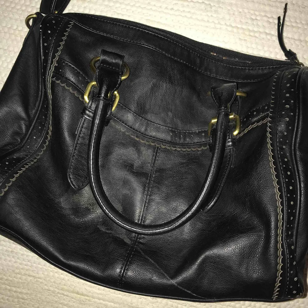 En hand- och axelremsväska i svart fake läder med gulddetaljer. Väskor.