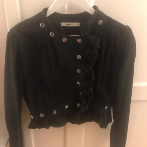  - Svart midjekort jacka från GinaTricot med volang detaljer vid knäppning och nedtill. Strl 38.