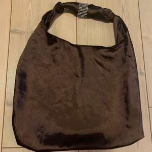 Tote bag  - En handsydd väska i brunt satin med en fin detalj i silver. Väskan är lite mindre, du får ex inte plats med din dator. Men perfekt att ha med ut på stan, på fest eller till vardags. Får plats med plånbok, telefon och läppstift ex. Även om du handlar mindre saker :) vid flera intresserade sker budgivning 