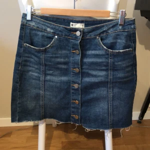 jeanskjol - en standard musthave jeanskjol med silvriga knappar och distressad kant i mörk tvätt, rymliga fickor. för stor i midjan för mig och skulle förmodligen passa någon i storlek 40 bättre om man vill att den ska sitta högmidjat!