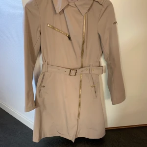 Beige Kappa Livelo - Beige kappa med gulddetaljer, från Livelo. Sparsamt använd! Skön vår/sommar-kappa då den är åt det tunnare hållet! Många fickor! Den är i storlek 36, men passar även storlek 38.