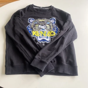 Svart sweatshirt från Kenzo med tigerbroderi - Säljer en helt ny svart sweatshirt från Kenzo med ett coolt broderi av en tiger i blått, gult och vitt på framsidan. Tröjan är långärmad och har en normal passform. Perfekt för både vardag och fest! 