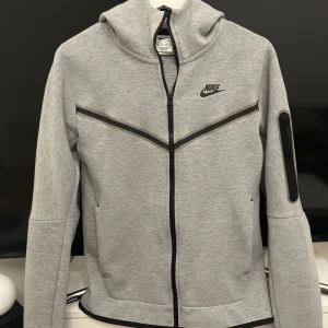 Grå hoodie från Nike - Säljer en snygg och bekväm grå hoodie från Nike. Den har en dragkedja framtill och en huva för extra mysfaktor. Perfekt för både träning och vardag. Den är långärmad och har en stilren design med svarta detaljer.