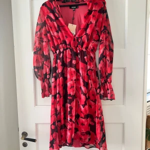 Röd blommig klänning från Missguided - Säljer en superfin röd klänning från Missguided med ett blommigt mönster i svart och rött. Klänningen har långa ärmar och en v-ringad design med resor i midjan. Perfekt för festliga tillfällen! Prislapp kvar och aldrig använd. 