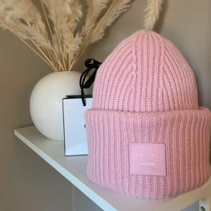 Rosa stickad mössa från Acne Studios - Säljer en superfin rosa stickad mössa från Acne Studios. Den har en ribbad design och ett ikoniskt ansiktsmärke framtill. Perfekt för att hålla sig varm och snygg under kyliga dagar. Passar till alla dina outfits!💖🩷💕