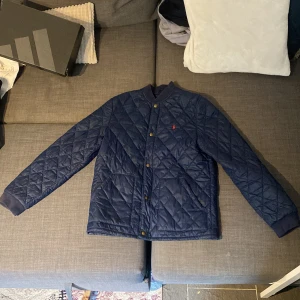 Blå quiltad jacka från Polo Ralph Lauren - Säljer en snygg blå quiltad jacka från Polo Ralph Lauren. Den har en klassisk design med knappar och en liten röd logga på bröstet. Perfekt för höst och vår, och den är långärmad för extra värme. Jackan är i bra skick och redo för nya äventyr!