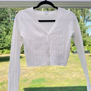 Croppad stickad tröja - En croppad vit ribbad tröja. Knappar framtill som är lite transparenta. Fin för sig själv med ett par högmidjade byxor eller öppen med en liten topp under 😍 Passar bra till mig som är en S/36.  (ALDRIG ANVÄND)  Längd: 43 cm Axel: 34 cm Byst: 80 cm