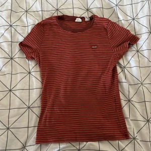 Röd- och vitrandig t-shirt från Levi's - Säljer en snygg röd t-shirt från Levi's med vita ränder. Den har en klassisk rund hals och korta ärmar. Perfekt för en avslappnad stil och passar till både jeans och shorts. Märket Levi's är broderat på bröstet. Skön och stilren för vardagsbruk!