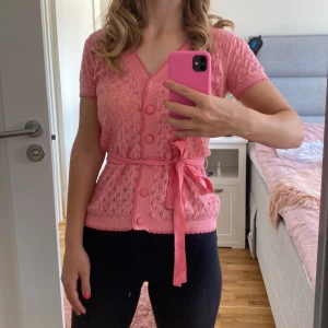 Rosa kofta med knytband - Säljer en superfin rosa kofta från Banned Apparel. Den har korta ärmar och ett snyggt knytband i midjan som ger en fin siluett. Koftan är stickad med ett vackert mönster och har knappar framtill. Perfekt för vår och sommar!