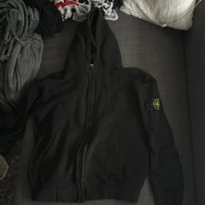 Svart hoodie från Stone Island - Säljer en svart hoodie från Stone Island med dragkedja och huva. Den har en ikonisk logga på ärmen. 