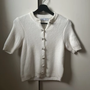 Vit kofta från Zara - Säljer en söt vit kofta från Zara med korta ärmar och dekorativa knappar. Perfekt för en stilren look. Koftan är mjuk och bekväm, idealisk för vår och sommar.