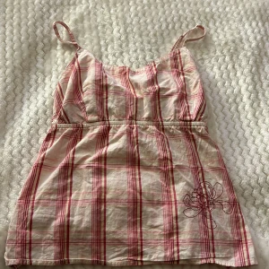 Rutig linne från H&M - Säljer ett somrigt linne från H&M med röd och vit rutigt mönster. Linnet har smala axelband och en broderad blomdetalj på framsidan. Perfekt för varma dagar!