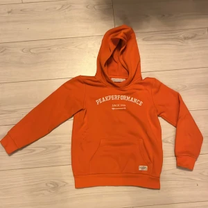 Barn: Orange hoodie från Peak Performance - Säljer en snygg klasisk orange hoodie från Peak Performance med vit text på bröstet, stl 140. Tröjan har en liten fläck vid höger ficka som säkert försvinner vid tvätt då den verkar uppkommit nu när vi ränsade ut lite kläder.