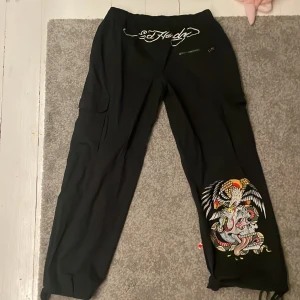 Svarta byxor från Ed Hardy - Snygga svarta byxor från Ed Hardy med coolt broderi av dödskalle på benet. Byxorna har flera fickor och dragkedja, vilket ger en praktisk touch. Perfekta för en unik och stilren look.