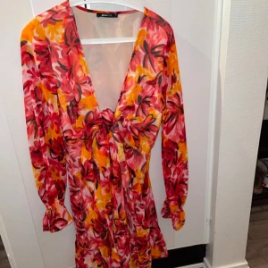 Blommig klänning från Gina Tricot - Färgglad blommig klänning från Gina Tricot med långa ärmar och v-ringad design. Klänningen har en knytning framtill och ett livfullt mönster i rött, rosa och gult. Perfekt för en somrig look!