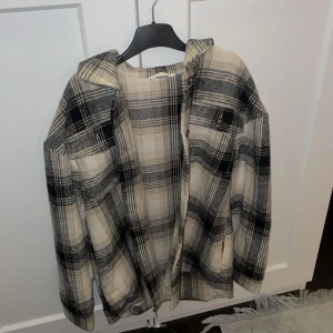 Rutig tunn jacka i beige och svart - Snygg och trendig jacka/skorta med rutigt mönster i beige och svart. Perfekt för lager-på-lager-stil. Den har knappar framtill och en avslappnad passform.