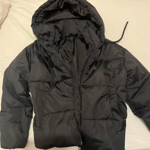 Svart pufferjacka med huva - Säljer en svart pufferjacka med huva och dragkedja. Jackan är perfekt för kyligare väder och har en bekväm passform. Den är tillverkad i ett syntetmaterial som håller dig varm och skyddad.