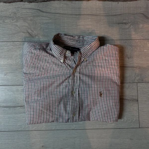 skjorta från Ralph Lauren - Säljer en klassisk rutig skjorta från Ralph Lauren i 100% bomull. Skjortan har en vit bas med röda och blå rutor samt knappar framtill. Perfekt för en stilren look.