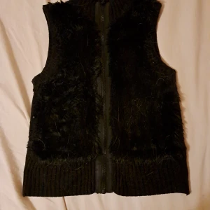 Svart pälsväst med dragkedja - Snygg svart väst med pälsdetaljer och dragkedja framtill. Perfekt för att hålla sig varm och stilren under kyligare dagar. Västen har en ribbad kant nedtill och vid ärmhålen.