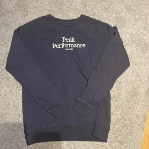 Mörkblå sweatshirt från Peak Performance - Säljer en stilig mörkblå sweatshirt från Peak Performance med vit text på bröstet. Tröjan har långa ärmar och ribbade muddar vid ärmslut och nederkant. Perfekt för en avslappnad stil.