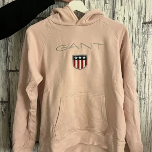 Rosa hoodie från GANT - Säljer en snygg rosa hoodie från GANT med ett broderat emblem och text på framsidan. Tröjan har en klassisk känguruficka och en bekväm huva. Perfekt för en avslappnad stil.