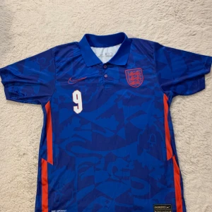 England borta vm2022 Kane 9 - Snygg blå fotbollströja från Nike med nummer 9 och namn på baksidan. Tröjan har korta ärmar och ett mönster i olika blå nyanser. Perfekt för fotbollsfans!