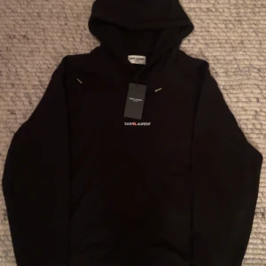 Svart Saint Laurent hoodie - Jag säljer en svarta Saint Laurent hoodie i nytt skicka med liten text. Hoodie är M som det står och passar runt 170-178cm. Den är i nytt skick och väldigt fresch. Väldigt snygg och matchar med många färger och klädesplagg🎉👍🏼