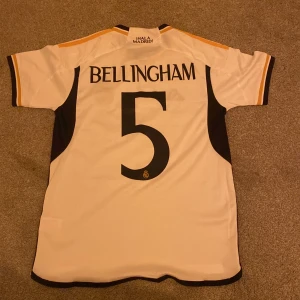  Real Madrid T-shirt - Säljer en vit Real Madrid fotbollströja från Adidas med Bellingham och nummer 5 på ryggen. Tröjan har svarta och gula detaljer samt klubbens emblem på bröstet. Perfekt för fans av laget! ⚽