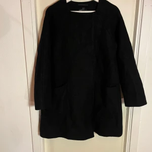 Svart kappa - Snygg svart kappa med långärmad design och enkel stil. Perfekt för kyligare dagar och ger en stilren look, 100% ull och håller värmen utan att den känns tung! Kappan har egentligen inga synliga defekter men är väl använd. Skicka ett meddelande om du undrar något!