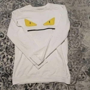 Vit tröja med gula ögon och dragkedja - Säljer en cool vit tröja med ett unikt tryck av gula ögon och en svart dragkedja på framsidan. Tröjan har långa ärmar och en avslappnad passform. Perfekt för en avslappnad stil med en twist! Fendi äkta pris kan diskuteras.