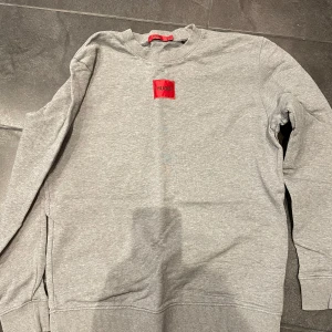 Grå sweatshirt från Hugo Boss - Säljer en stilren grå sweatshirt från Hugo Boss med röd logga på bröstet. Tröjan har rund hals och långa ärmar, perfekt för en avslappnad look. Passar bra till både jeans och joggers.