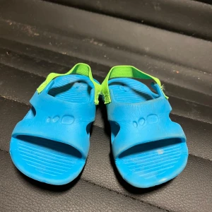 Blåa bad sandaler  med gröna detaljer - Säljer ett par blå sandaler med gröna elastiska remmar. Perfekta för sommarens äventyr och bad. Sandalerna har en enkel design med öppen tå och är tillverkade i syntetmaterial.