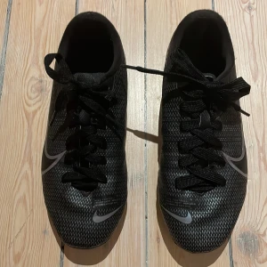 Svarta Nike Mercurial fotbollsskor  - Säljer ett par svarta Nike Mercurial fotbollsskor med snörning och multiground dobbar för bra grepp på planen. Sulan har gått upp lite på kanten men är limad med vattentätt lim och fungerar som vanligt. Skorna har mönster och Nike-logga i silver. Perfekta för fotbollsträning och matcher.