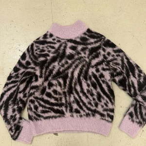 Lila  och svart mönstrad tröja - Säljer en fluffig och mysig tröja i rosa och svart med ett djärvt zebramönster. Tröjan har långa ärmar och en ribbad kant vid halsen, ärmsluten och nederkanten. Perfekt för att sticka ut i mängden!