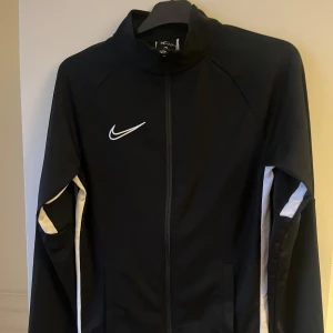 Svart träningsjacka från Nike - Snygg svart träningsjacka från Nike med Dri-FIT-teknologi. Jackan har en dragkedja framtill och vita detaljer längs ärmarna. Perfekt för träning och sportiga aktiviteter. Står XS i tröjan men uppskattar att det är en S.