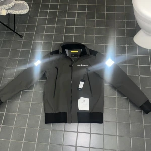 Grå jacka från Sail Racing - Snygg grå jacka från Sail Racing med svarta detaljer och dragkedja. Jackan har reflexdetaljer på ärmarna och en logotyp på bröstet. Perfekt för en sportig look. Fick den i julklapp men var för liten . Så de kan kanske bli en ny julklapp …. Köpt för 2800kr passar både s och m väldigt slim fit 