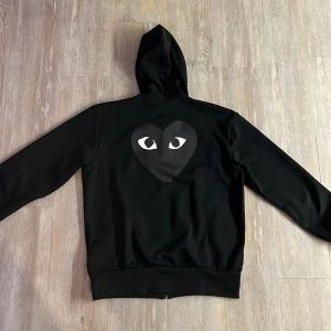 Svart hoodie från Comme des Garçons - Säljer en svart hoodie från Comme des Garçons med det ikoniska hjärtmotivet med ögon på bröstet och ryggen. Hoodien har dragkedja och långa ärmar, perfekt för en stilren look. Nypris 3000kr.