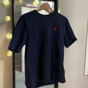 Ami paris t-shirt - Snygg mörkblå t-shirt från Ami Paris med en liten röd broderad detalj på bröstet. Perfekt för en stilren och avslappnad look. Kortärmad och tillverkad i mjukt material. Tirsan är inprencip helt ny. Vid frågor och annat finns jag alltid i dm!