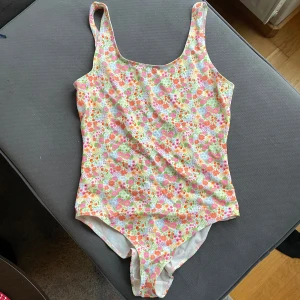 Blommig baddräkt Monki - Oanvänd snygg baddräkt med färgglatt blommönster i rosa, orange och grönt på vit bakgrund. Perfekt för sommarens badäventyr!