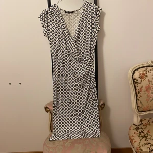 Mönstrad klänning från Esprit - Snygg klänning från Esprit med ett geometriskt mönster i blått och vitt. Klänningen har en omlott-design och korta ärmar, vilket ger en stilren look. Perfekt för både vardag och speciella tillfällen.