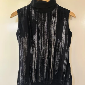 Svart ärmlös polotopp med silvermönster - Snygg svart ärmlös polotopp med vertikalt silvermönster. Perfekt för en stilren look med en touch av glamour. Polokragen ger en elegant känsla.🌟