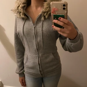 Grå hoodie med päls - Mysig grå hoodie med dragkedja och fuskpäls på luvan. Perfekt för kyliga dagar med sina långa ärmar och bekväma passform. Praktiska fickor framtill för extra värme och stil.  Det är mina bilder!