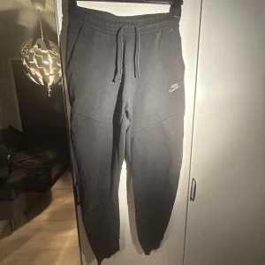 Svarta Nike tech byxor från Nike, använt skick - Bra pris för svarta Nike tech byxor!! Dem är i använt skick där man ser att ens snörets metallbit gåt av och tyget på baken är lite slitet fast de är inte märkbart