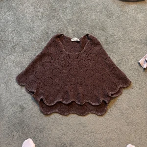 Brun poncho från Mille K - Säljer en brun poncho från Mille K med ett vackert spetsmönster. Ponchon har en vågig kant och är perfekt för lager-på-lager-stil. Ett unikt plagg för att lyfta din garderob!