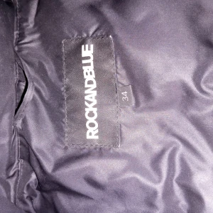 Svart dunjacka från Rockandblue - Snygg svart dunjacka från Rockandblue med quiltad design och pälskantad huva. Jackan har dragkedja och knappar framtill samt praktiska fickor med dragkedja. Perfekt för kyliga dagar.